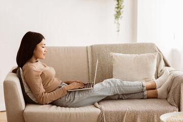 Obraz premium A young woman relaxes on a couch with her feet up, using a laptop. She is wearing casual clothing and appears to be working or browsing the internet.