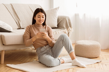 A young woman sits on a rug in a living room, using a smartphone. She is wearing a beige sweater and blue jeans.