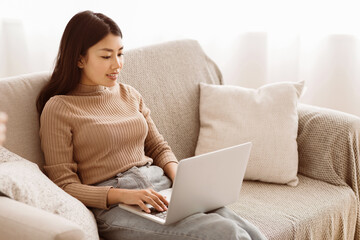 A young woman sits on a sofa in a home setting, working on a laptop.