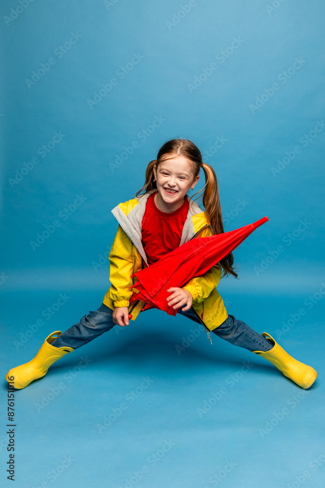 Wall mural Happy little emotional cheerful child girl laughing, with red umbrella posing on blue wall background. Girl is wearing yellow raincoat and rubber boots. Childhood, copy space