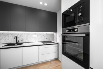 A modern kitchen with black and white cabinets, a terrazzo countertop and backsplash, a black sink, an electric cooktop, and built-in black oven and microwave.
