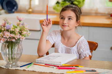 a girl draws in an album with colored pencils while sitting at a table in the interior of a country house kitchen.