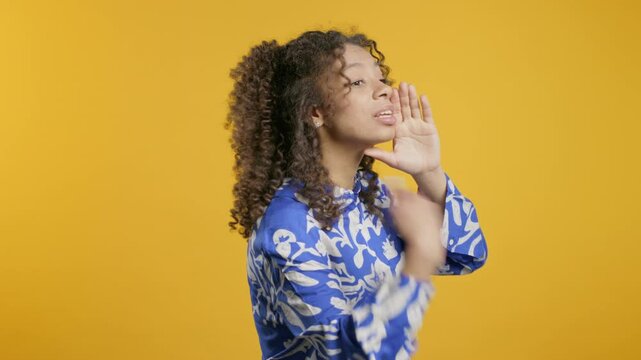 Attention, portrait of young african american girl shouting, calling, holding hand near open mouth isolated on yellow background with copy space for your advertisement
