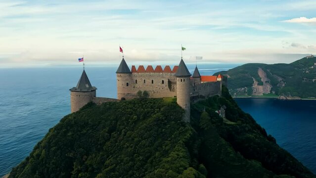 Aerial shot. The camera captures the expansive castle situated on a rocky island surrounded by azure waters. Waves gently lap against the base of the island, emphasizing its isolation and grandeur.