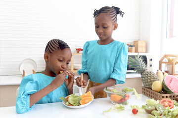 Twin sister kid girl with curly hair braid African eating fresh vegetables and fruits after washing and cleaning in glass bowl for making salad. Cute children preparing salad healthy food at kitchen.