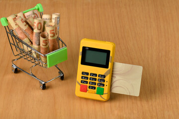 a yellow payment machine with a beige debit card and a shopping cart full of Brazilian money