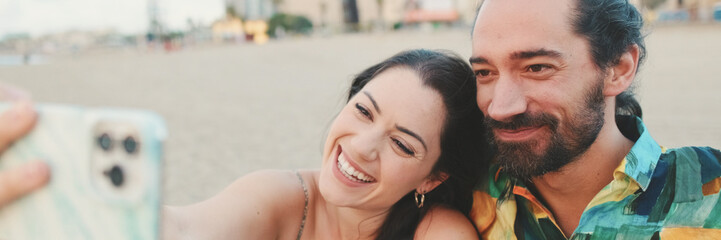 Happy couple taking selfie on mobile phone while sitting on the beach, panorama
