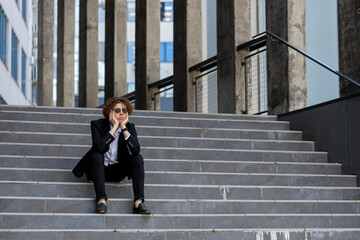 Sad Caucasian young businessman sitting outside on the steps of an office building. Male in a formal suit holds his hand on his head. Corporate employee with curly hair went bankrupt or lost his job.