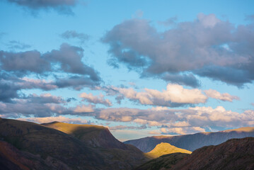 Mountain and hill tops illuminated by sunset light. Rocks silhouettes and gold sunrise colors. Vivid golden sunlight on hills under clouds of sunset tones in blue sky. Shadows of clouds on rocky ridge