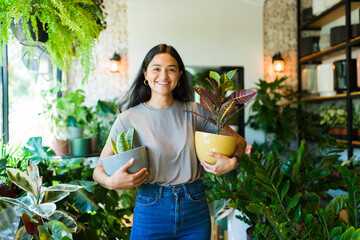 Portrait of a joyful young woman shopping for houseplants at a garden center, holding two potted...