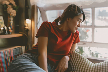 Sleepy asian woman in red t-shirt resting in the living room near the windows.