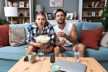 Young couple, man and woman, sitting at home on sofa, eating popcorn and watching scary horror movie or watch dramatic sport game action or drama film. Simple living concept, weekend activities.