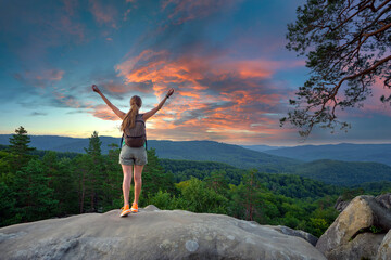 Sportive woman standing alone on hillside trail with raised up arms. Female hiker enjoying view of evening nature from rocky cliff on wilderness path. Active lifestyle concept © bilanol
