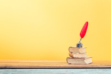 Vintage red quill pen with inkwell and old books on oak wooden desk front yellow background. Retro style filtered photo