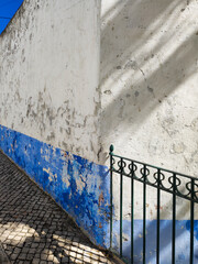 Narrow alley with a weathered wall and green iron fence. White and blue old wall with peeling paint