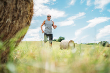 Smiling man jogs by scenic countryside hay bales in background, embodying essence of trail running and ultramarathon training. Clear sky and lush landscape highlight harmony of outdoor fitness nature.