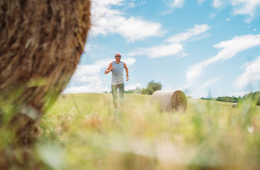 Smiling man jogs by scenic countryside hay bales in background, embodying essence of trail running and ultramarathon training. Clear sky and lush landscape highlight harmony of outdoor fitness nature.