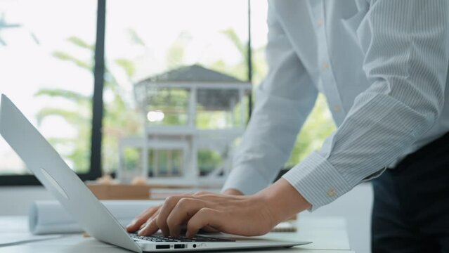Close up of professional male architect engineer measure house model and using laptop analysis data at meeting table with house model and map . scatter around. Focus on hand. Immaculate.
