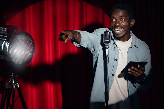 Smiling African American man speaking to microphone performing in comedy show open mike stand up on stage with red curtains.