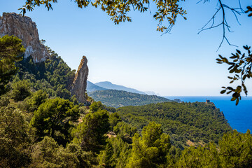 Monolith Es Penyal Bernat framed by lush vegetation and historic Torre Picada tower, Mediterranean Sea in the background, captured on a sunny spring day, perfect Mallorca coastal hiking destination.