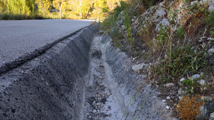 A concrete drainage ditch runs parallel to a winding road, bordered by a rocky hillside. Wild...