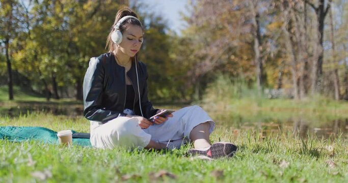 Beautiful girl listening to music on a meadow in the park. Fashionable young lady puttion on headphones connected to her phone.