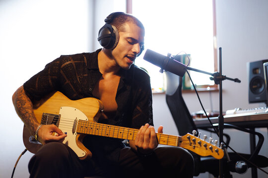 Stylish mixed race man in headphones playing electric guitar near microphone in studio