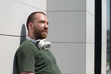Fototapeta premium Portrait of caucasian handsome beard man wearing military color t-shirt with headphones near modern white wall listening music. Favourite song. Copy space.