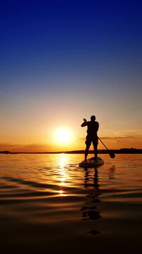 Practicing paddle boarding on the calm river in summer. Male silhouette stands on the sup board rowing with an oar. Low angle view. Vertical video.