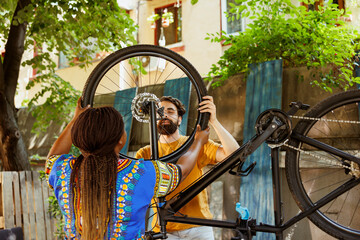 Young multiethnic couple happily attaching new bicycle tire for outdoor summer cycling. Black woman...
