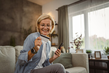 Obraz premium Mature senior woman buy online on mobile and use credit card on sofa