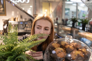 A young woman with long red hair leans over a glass case, her eyes fixed on the delectable pastries within. She appears to be lost in thought, perhaps imagining the sweet taste of the treats.