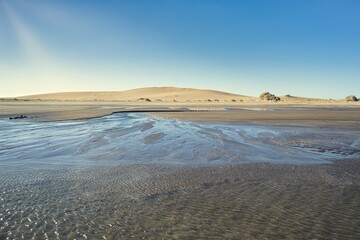 tranquil beach scene featuring rippled wet sand that reflects the sunlight.Background with large dry sand dunes, clear blue sky and sun rays