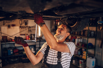 Smiling professional mechanic under lifted car fixing malfunction.