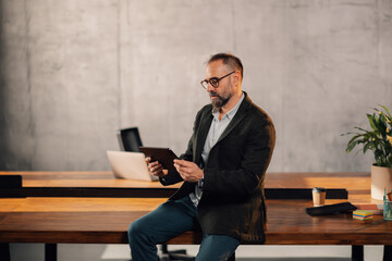 Middle aged manager sitting on table at modern office and using tablet