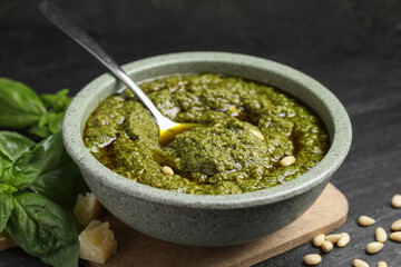 Tasty pesto sauce in bowl, pine nuts, cheese and basil on black table, closeup