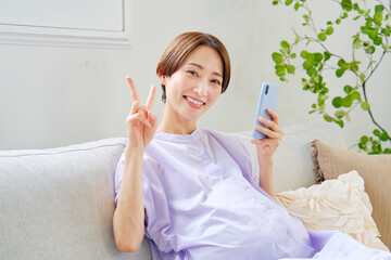 自宅でスマートフォンを持ってピースサインをする高廉妊娠をした妊婦