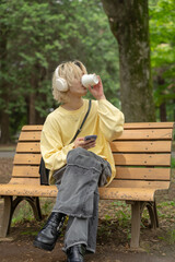 In Yoyogi Park, Shibuya, Tokyo, a Japanese man in his twenties wearing yellow clothes, sitting on a secluded bench, operating a smartphone along with headphones, music, and coffee.