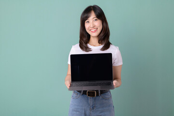 Technology advertisement. cheerful beautiful Asian woman in white t-shirt and holding laptop mockup of blank screen on green background.