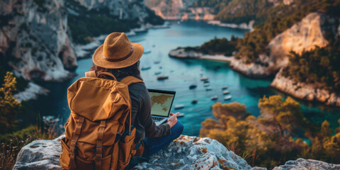 A traveler wearing a hat and backpack uses a laptop while sitting on a cliff, overlooking a stunning bay with boats.