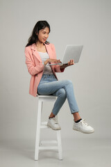 full lenght shoot of happy asian woman holding laptop computer sitting on stool on isolated background