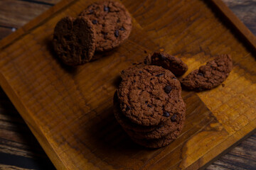 Brown chocolate chip cookies are placed on a brown wooden tray with a glass of milk and a brick wall in the background.