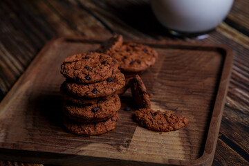 Brown chocolate chip cookies are placed on a brown wooden tray with a glass of milk and a brick wall in the background.