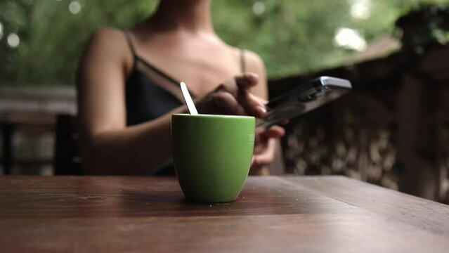 Middle-aged Woman sitting at a table, drink green tea and texting on the smartphone. Online Chat During Breakfast on Exotic resort against green, blured forest background.