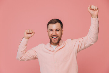 Cute unshaven man in pink shirt waving fists in joy smiling and posing isolated on pink background...
