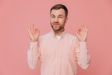 Cute unshaven man in pink shirt showing okay with hands isolated on pink background with copy space
