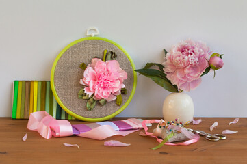 Handmade embroidery with satin ribbons of peony flower in hoop, bouquet of fresh peonies in vase on wooden table. Nearby is set of colorful satin ribbons, needles and scissors for work. DIY concept