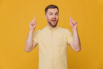 Cute unshaven man in yellow shirt pointing fingers up at copy space isolated on yellow background