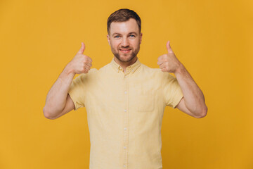 Cute unshaven man in yellow shirt showing thumbs up isolated on yellow background