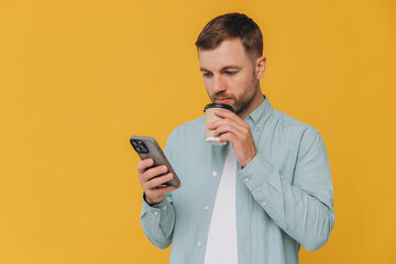 Unshaven man holding smartphone in one hand and takeaway coffee cup in another, looking at screen of phone, isolated on yellow background